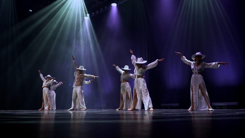 Cinq danseurs en blanc et chapeaux, sous des faisceaux de lumière.